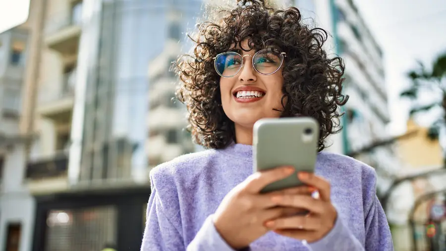 Woman standing smiling with her smartphone in hand in front of a building